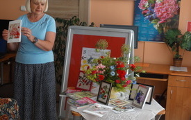 Kobieta w średnim wieku prezentuje publiczności oprawiony w ramkę obraz z kwiatami