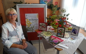 Starsza kobieta w okolurach siedzi przy stoliku, na kt&oacute;rym stoją kwiatowe dekoracje