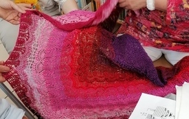 Trzy kobiety rozkładają dużą kolorową chustę zrobioną na szydełku
