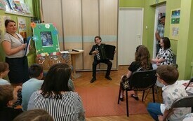 Dzieci i dorośli siedzą w bibliotece słuchając recitalu chłopca grającego na akordeonie