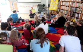 Dzieci wraz z rodzicami siedzą na podłodze w bibliotece oglądając książkę prezentowaną przez bibliotekarkę