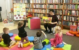 Bibliotekarka pokazuje siedzącym na podłodze dzieciom ksiażkę z obrazkami
