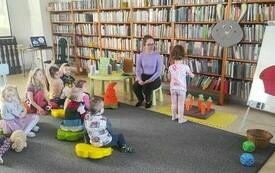 Młoda kobieta siedzi z grupą dzieci, w tle regały biblioteczne