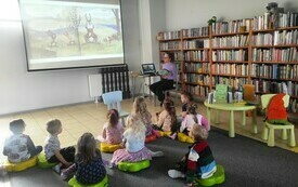 Dzieci siedzą na podłodze w bibliotece i oglądają bajkowe ilustracje na ekranie