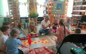 Na dywanie w bibliotece grupa dzieci. Przed dziećmi na podłodze żyrafa wykonana z kolorowego papieru.