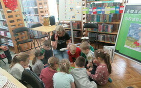 Na podłodze w bibliotece grupa dzieci wraz z bibliotekarką.