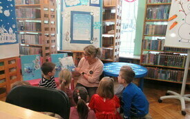 Kobieta pokazuje dzieciom książkę w tle regały biblioteczne.