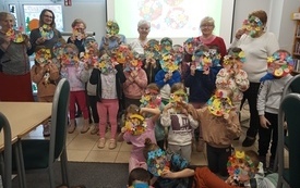 Grupa dzieci i seniorek na zdjęciu grupowym. Trzymają w ręku prace plastyczne z papieru.