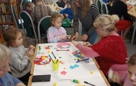 Stół z materiałami plastycznymi, wokół niego babcie i wnuki. W tle więcej osób pracujących przy stołach.