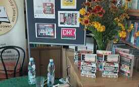 Książki na stoliku, po prawej stronie bukiet kwiat&oacute;w. W tle na tablicy logo DKK. 