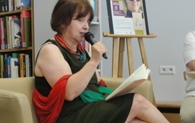 Kobieta w sukience, koralach i szalu siedzi na fotelu i czyta do mikrofony.