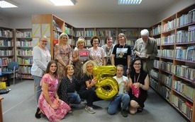 Grupa ludzi stoi i siedzi obok siebie, kobieta trzyma balon z cyfrą 5. W tle regały z książkami.
