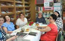Grupa kobiet siedzi przy stole, wok&oacute;ł regały z książkami. 