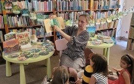 W tle regał z książkami, na środku bibliotekarka z książką, a przed nią dzieci. 