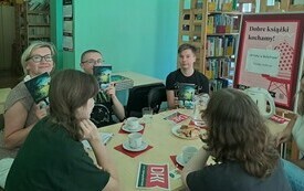 Grupa młodych ludzi siedzi przy stole, w tle regały z książkami.