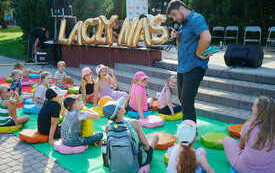 Mężczyzna z mikrofonem stoi pośród siedzących na dywanie dzieci.