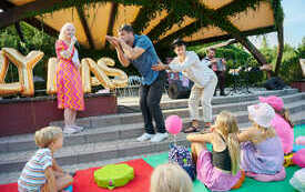 Kobieta w różowo-pomarańczowej sukience z mikrofonem w ręku, obok dwóch młodych mężczyzn pokazuje coś publiczności.