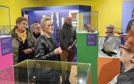 Przewodniczka muzealna stoi pośrodku grupy senior&oacute;w zwiedzajacych muzeum. Dookoła gabloty z zabytkowymi butami