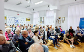 Publiczność zebrana na spotkaniu siedzi na krzesełkach w sali.