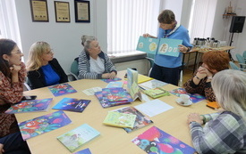 Pięć kobiet w średnim wieku siedzi przy stole wypełnionym książkami i patrzy na prowadzącą, która prezentuje jedną z książek