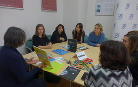 Siedem kobiet w średnim wieku siedzi przy stole i patrzy na prowadzącą, która prezentuje książkę