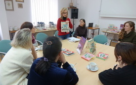 Sześć kobiet w średnim wieku siedzi przy stole i patrzy na prowadzącą, która prezentuje książkę