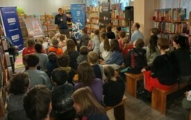 Podróżnik przemawia do dzieci w otoczeniu regałów z książkami.