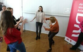 Kilka kobiet trzyma ręce przed sobą, jedna z nich stoi, pozostałe pokazują na nią.