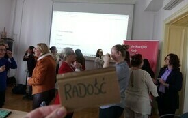 Na pierwszym planie ręka trzyma kartonik z napisem "Radość", w tle grupa bibliotekarzy pokazuje miny i odgrywa dramowesceny.