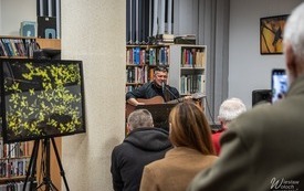 Muzyk śpiewający i grający na gitarze. Na sztaludze fotografia gałęzi drzewa.