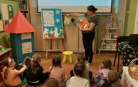 Grupa dzieci  siedzi na podłodze, przed nimi bibliotekarka prezentuje książki o Muminkach