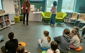 Grupa dzieci siedzi na podłodze w bibliotece i ogląda prezentację na ekranie