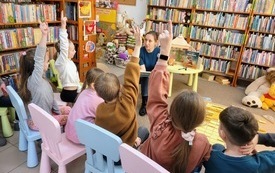 Grupa dzieci siedzi na krzesełkach i słucha książki czytanej przez bibliotekarkę
