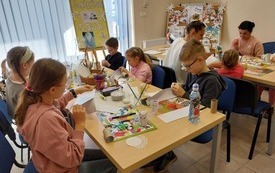 Grupa dzieci siedzi przy stolikach wykonując prace plastyczne