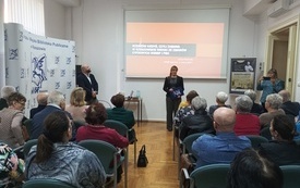 W sali wypełnionej widownią - bibliotekarka wita gościa na spotkaniu.