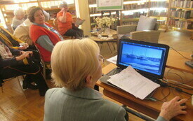 Kobieta przed komputerem, z boku uczestnicy słuchający prelekcji.