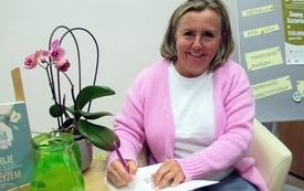 Kobieta w różowym sweterku siedzi przy stoliku i podpisuje autografy.