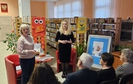 Dwie kobiety stoją przed zgromadzona publicznością, w tle regały z książkami.