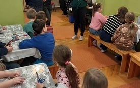 Grupa dzieci siedząca przy stolikach, dzieci zwr&oacute;cone w kierunku prowadzących spotkanie.