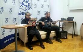 Na sofie mężczyzna grający na gitarze i kobieta czytająca teks do mikrofonu.