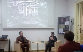 Mężczyzna i kobieta siedzą przy stoliku w sali, w tle prezentacja multimedialna.