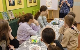 Młodzież siedzi przy stole na którym znajdują się materiały plastyczne. Obok stoi kobieta.