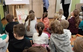 Grupa dzieci siedzących na dywanie, przed nimi kobieta trzymająca w ręku papierowego kr&oacute;lika i dziewczynka.