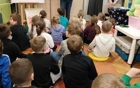 Trzy kobiety stojące przed grupą siedzących na dywanie dzieci.