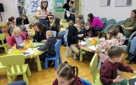 Grupa dzieci z opiekunami siedzi przy stolikach w sali bibliotecznej i wykonuje prace plastyczne