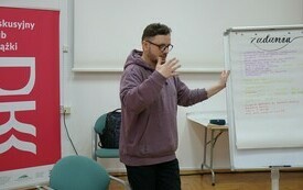 Mężczyzna w bluzie i okularach tłumaczący coś zebranym. 