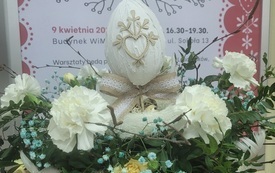 Dłonie trzymające stroik świąteczny na tle plakatu z wydarzenia.