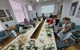 Duży stół z materiałami florystycznymi, przy którym siedzą kobiety w różnym wieku.