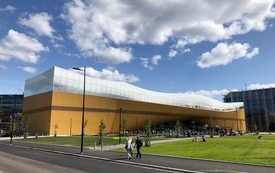 Długi żółty budynek z falującym szklanym dachem, wokół zielone trawniki