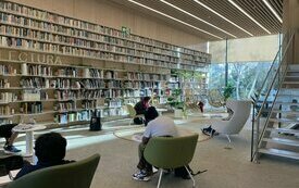 Wnętrze biblioteki z wysokimi do sufitu półkami z książkami, obok nich czytelnicy w fotelach